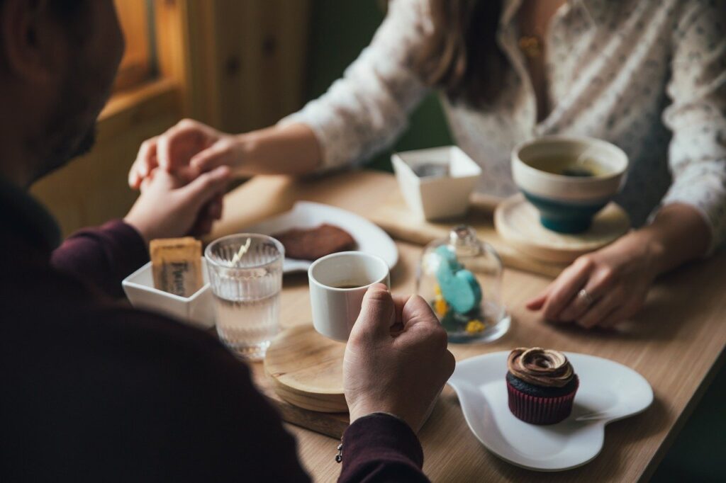Hochzeitsplanung Stück für Stück feiern: Ein paar hält sich an den Händen, während es an einem Tisch mit Kaffee, Törtchen & Co. sitzt. 